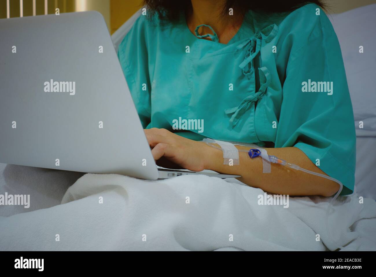 A half body shot of a workaholic business woman sitting on her hospital bed with IV injection in her arm, recovering from surgery while using her lapt Stock Photo