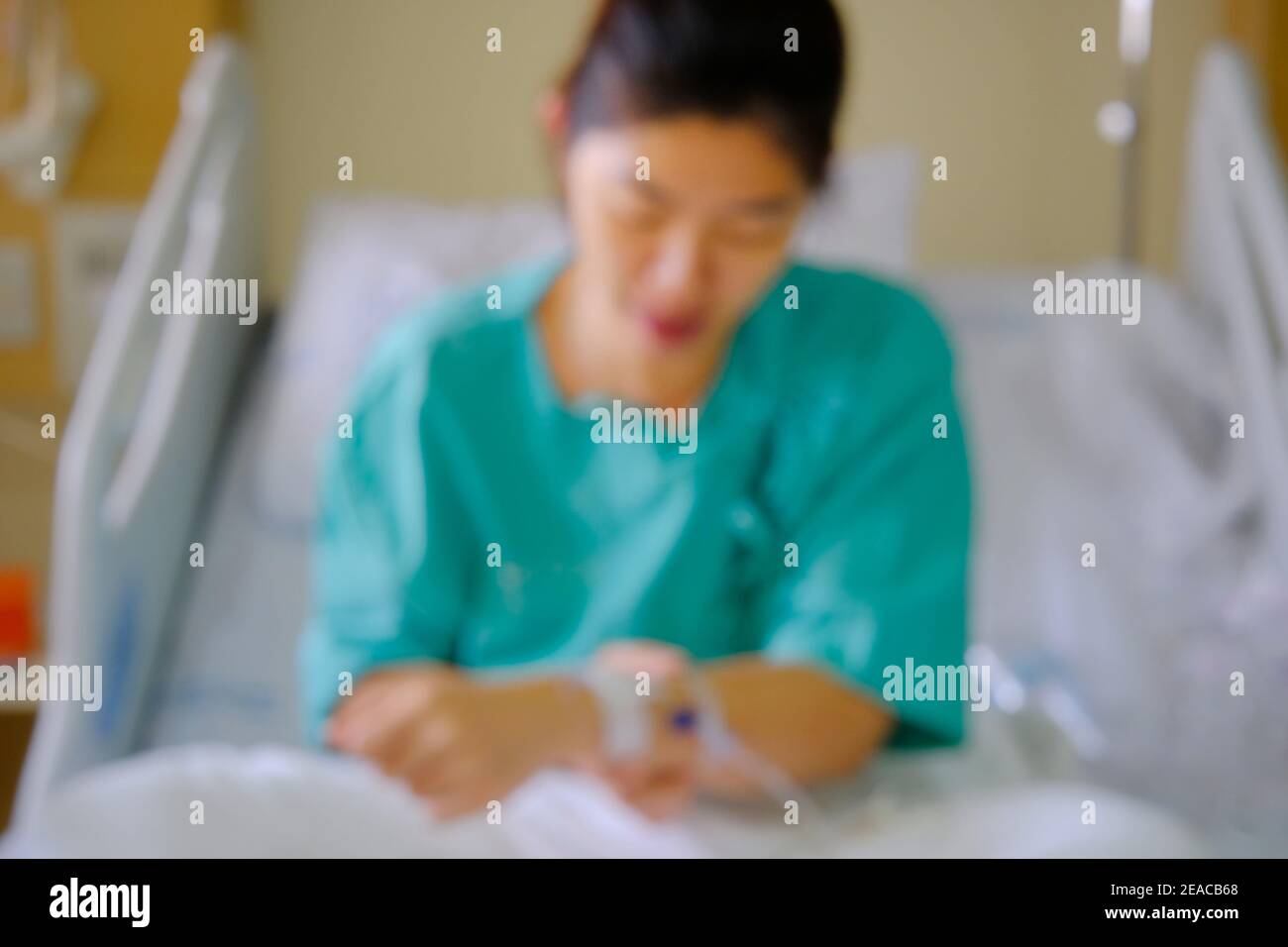 An out of focus picture of an Asian woman being treated for her illness in a hospital room, sitting on a bed with a green uniform and saline solution Stock Photo