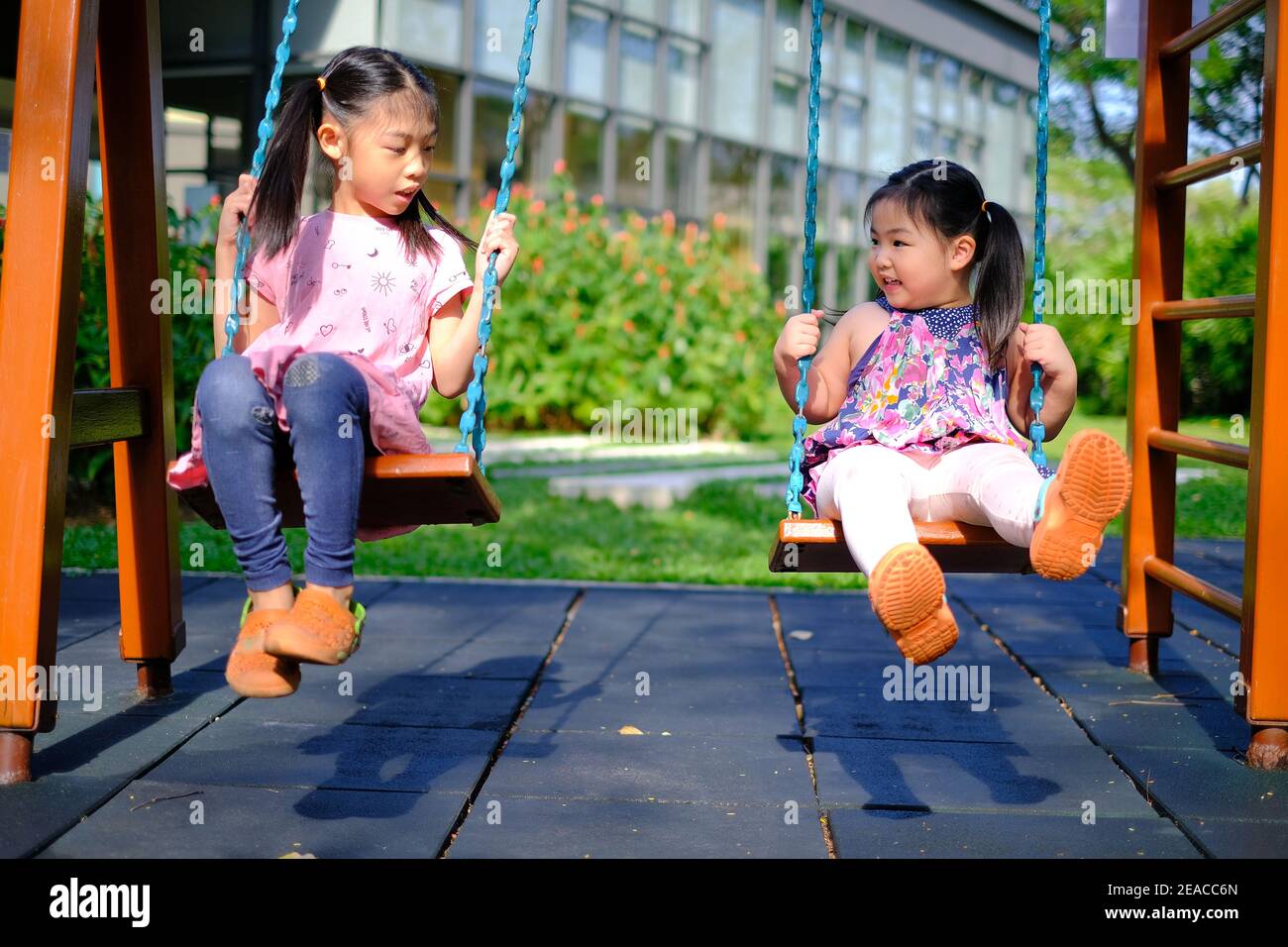 Two cute young Asian girls at a playground playing swings together, having fun outdoor and smiling on a sunny day. Stock Photo