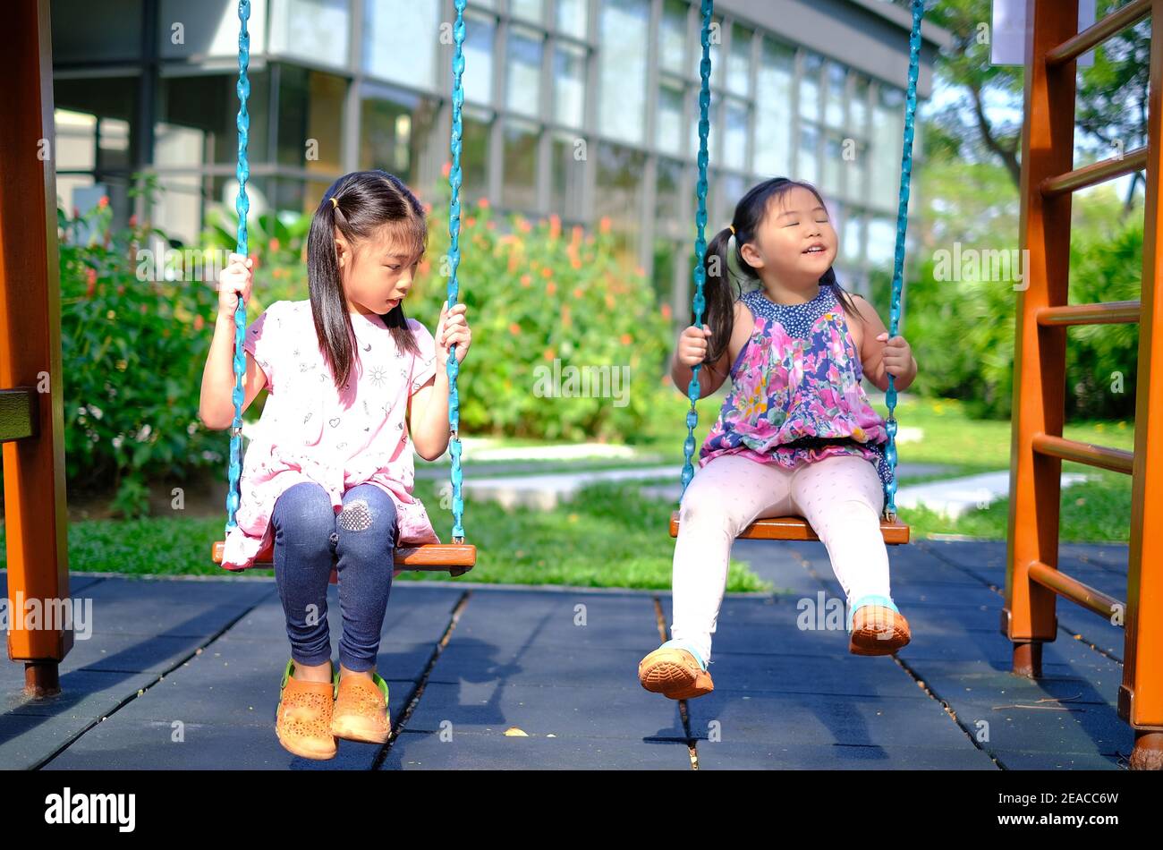 Two cute young Asian girls at a playground playing swings together, having fun outdoor and smiling on a sunny day. Stock Photo