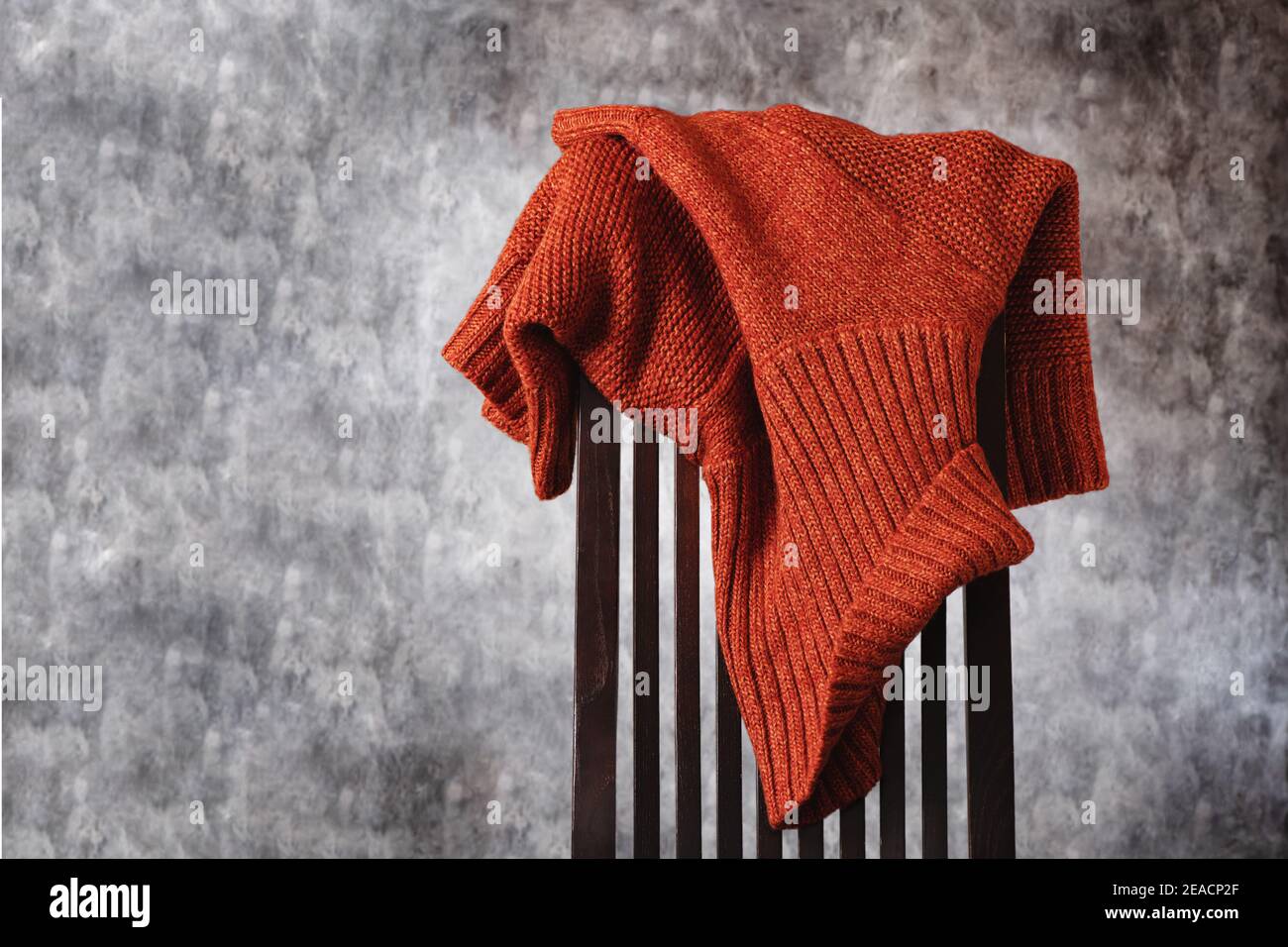 Women's wool knitted terracotta sweater casually thrown over the back of wooden chair against gray wall. Minimal style. Copy space Stock Photo