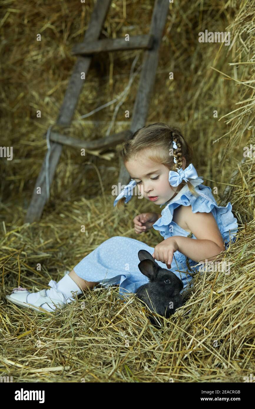 Little pretty girl is sitting in the hay with a little rabbit. Girl is feeding her favourite pet. Summer in a village. Happy childhood Stock Photo