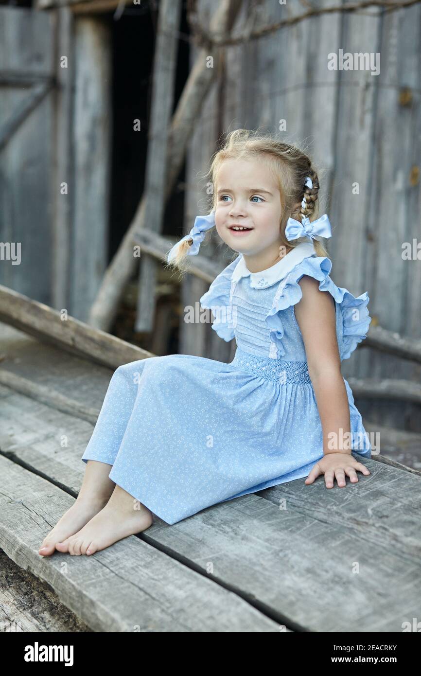 Cute little funny girl in a blue dress is sitting in the village yard ...