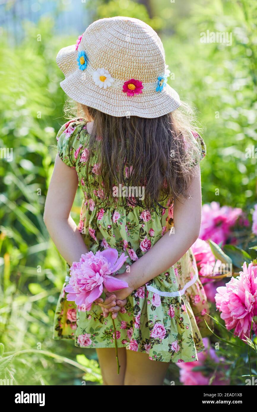 Beautiful little girl is holding a pink peony in a sunny day in the garden.  Pretty girl is wearing a fashionable patterned summer dress and straw hat Stock Photo