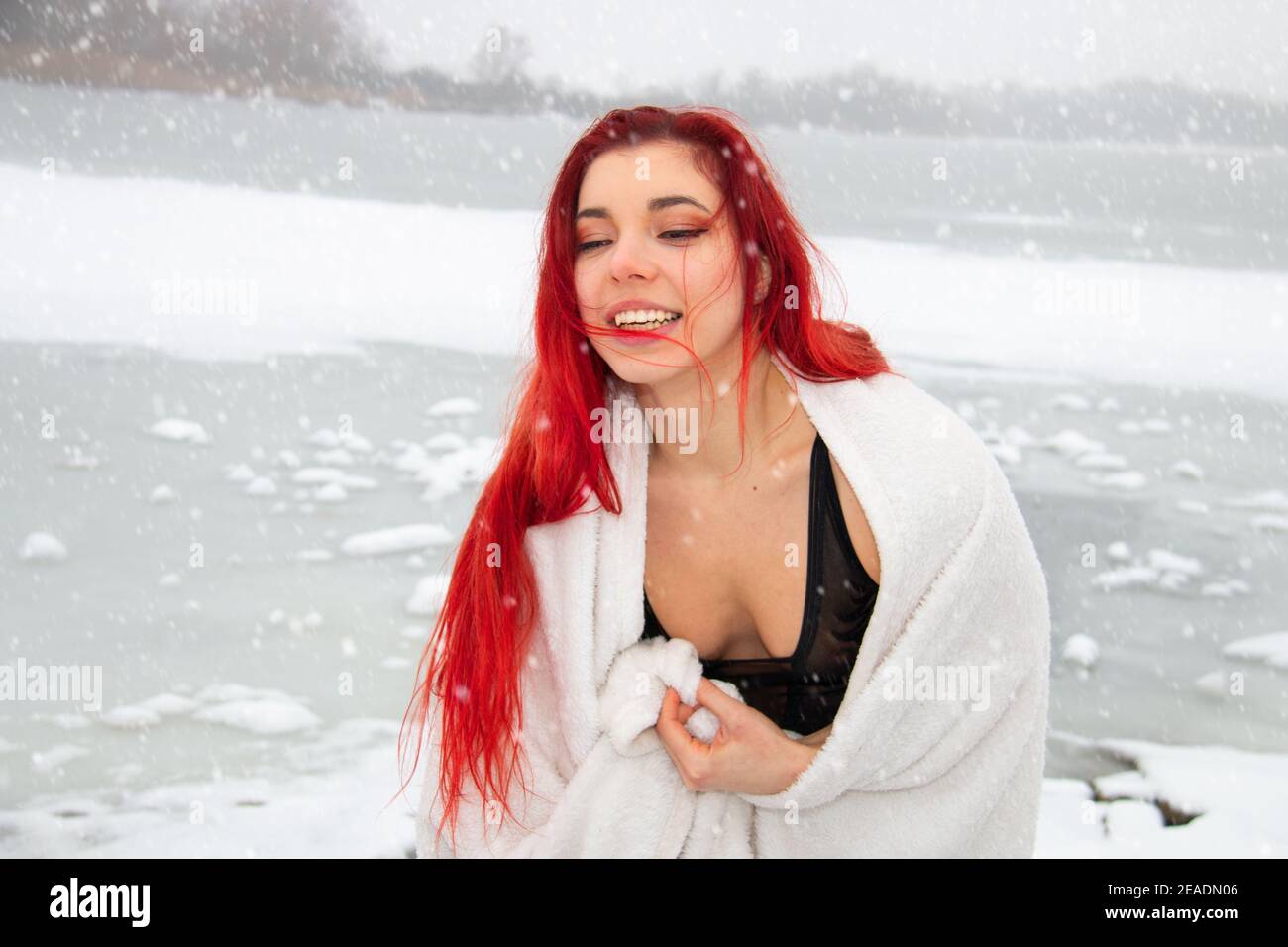 Snowy winter portrait of red hair woman outdoors in a bathing suit, wrapped in a towel at open water swimming lake, cold therapy and hardening on snow Stock Photo