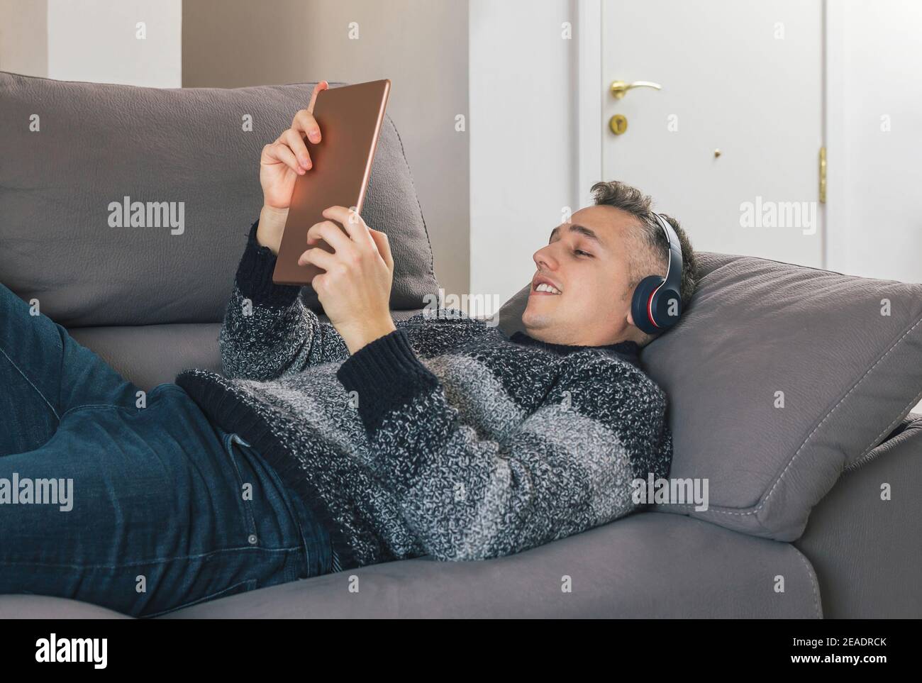 Young man lying and relaxing on the sofa while listening music and using a tablet. Guy having great time at home concept Stock Photo