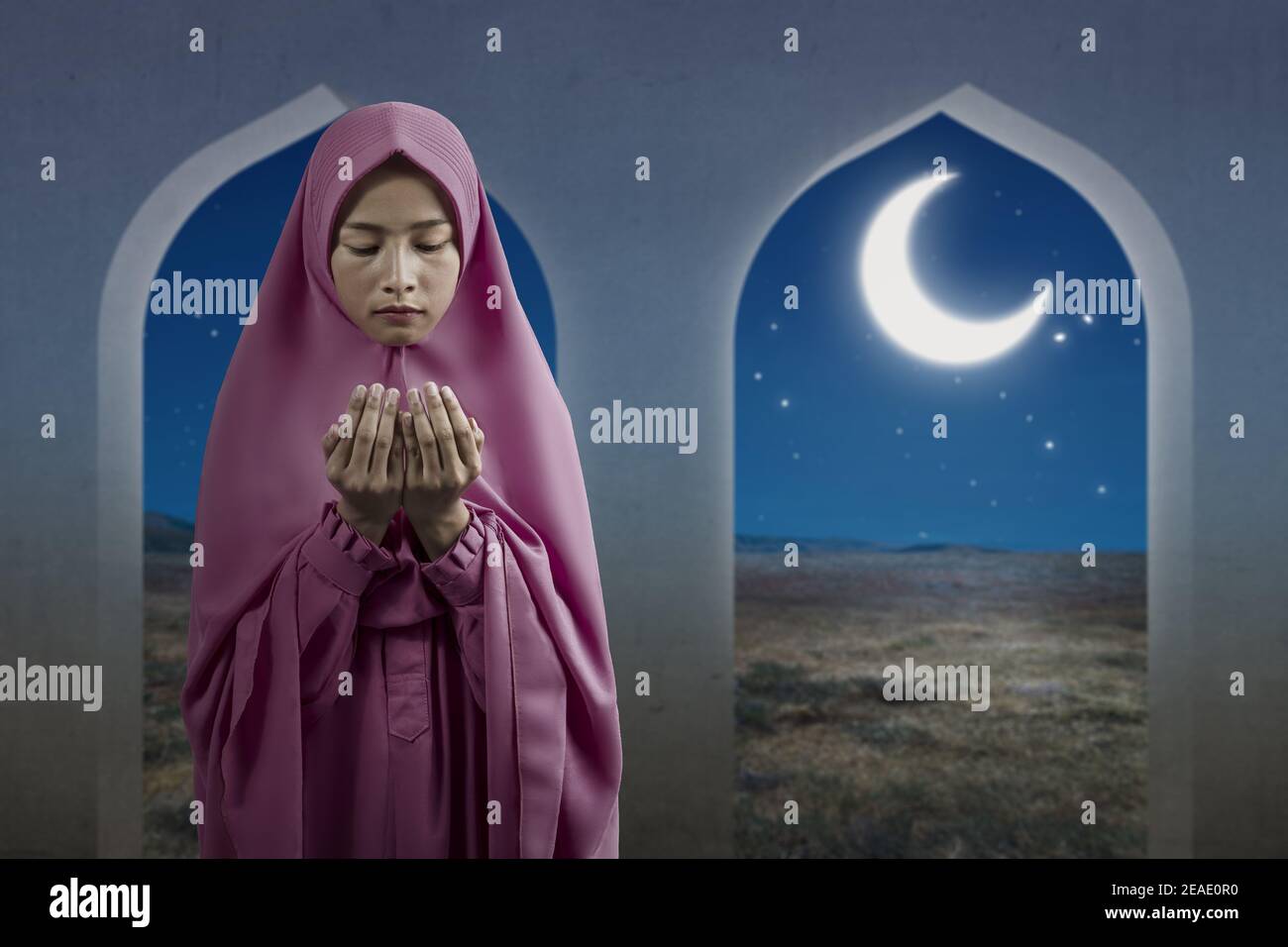 Asian Muslim woman in a veil standing while raised hands and praying on ...