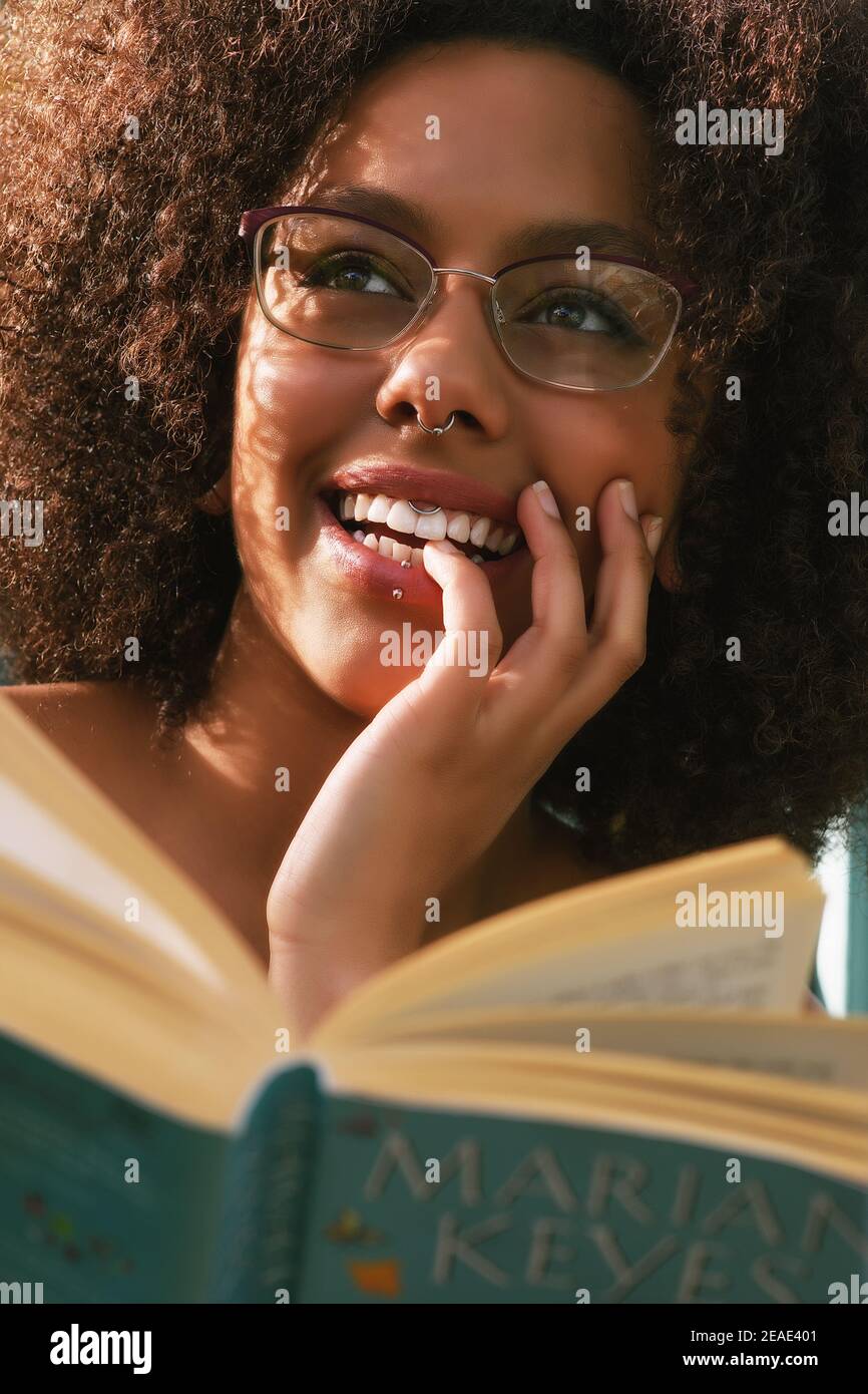 Stunning black Female model for editorial photo shoot, wearing glasses and smiling, reading a book outside in natural sun light Stock Photo