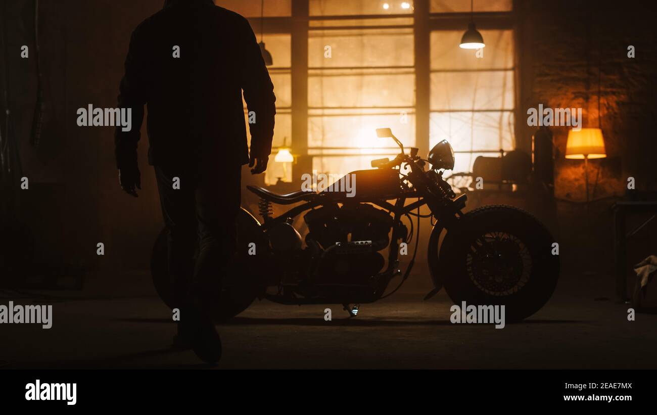 Custom Bobber Motorbike Standing in an Authentic Creative Workshop. Silhouette of a Rider Coming to a Bike. Vintage Style Motorcycle Under Warm Lamp Stock Photo