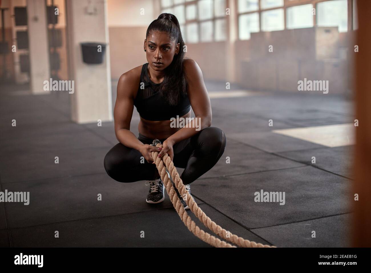 Fit young Indian woman in sportswear crouching on a gym floor and looking exhausted after a battle rope workout session Stock Photo