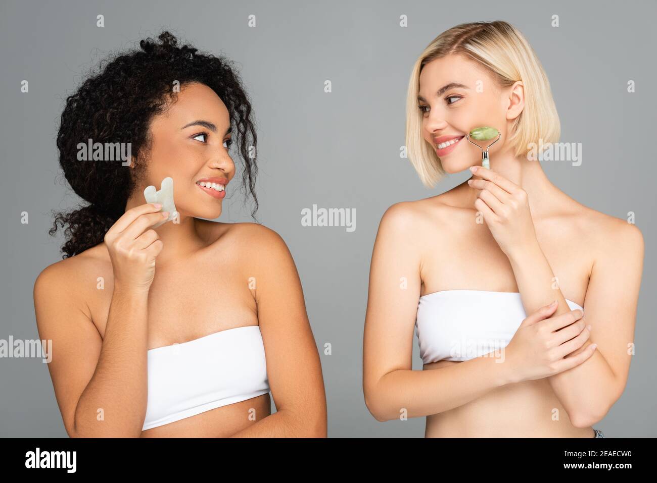 Smiling multiethnic women in white tops using jade roller and gua sha isolated on grey Stock Photo