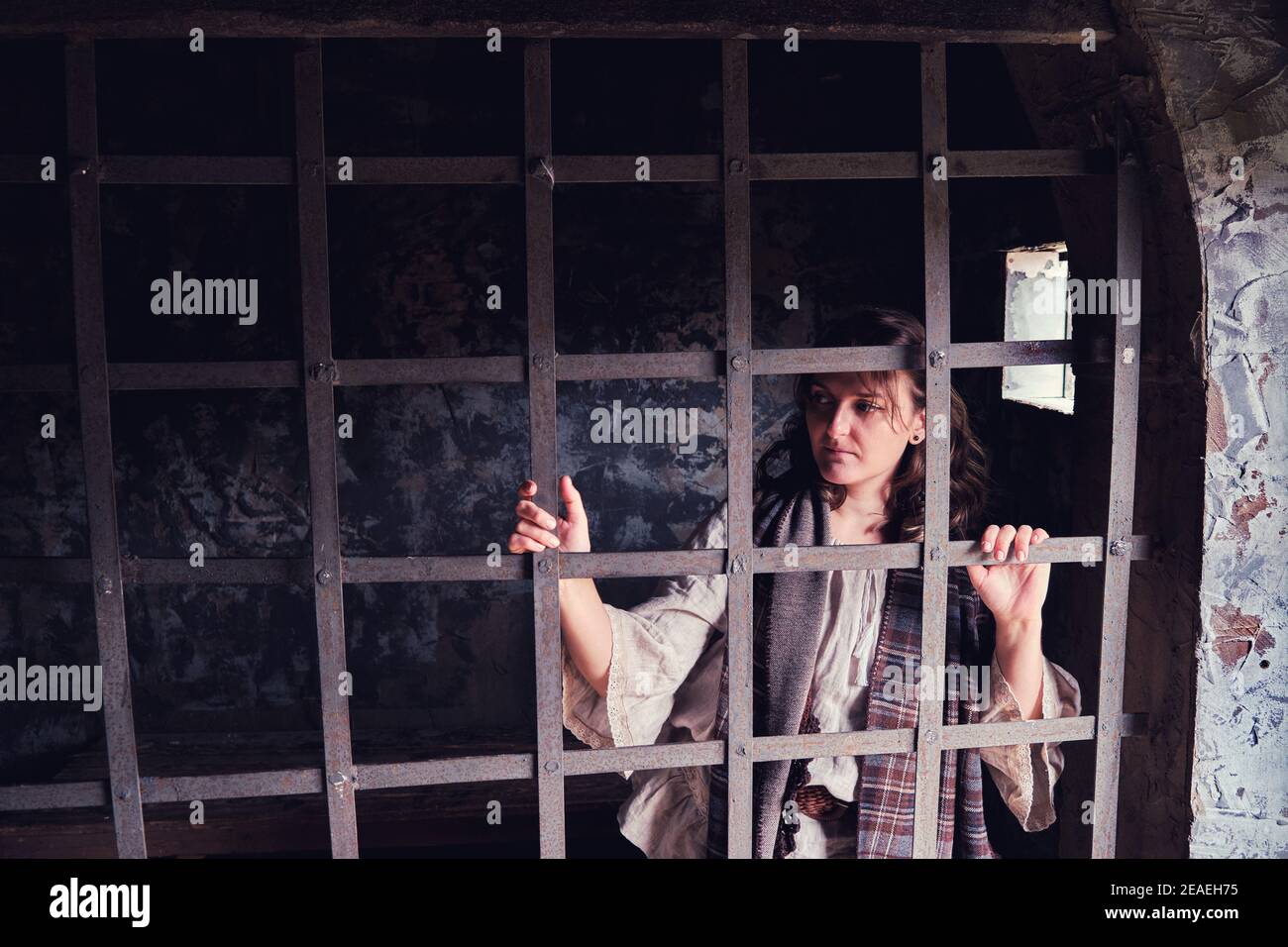 A woman looks through the bars of an old-style prison cell. A young woman in a vintage dress from the 18-19 century. Stock Photo