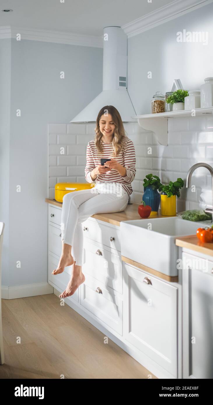 Beautiful Young Female in Striped Jumper and White Pants is Sitting on a Kitchen Furniture and Using Her Smartphone in a Modern Sunny Kitchen. She is Stock Photo
