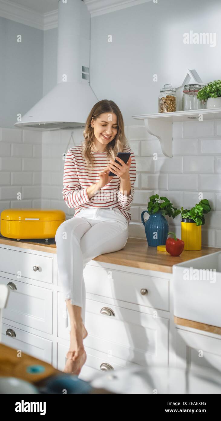 Beautiful Young Female in Striped Jumper and White Pants is Sitting on a Kitchen Furniture and Using Her Smartphone a Modern Sunny Kitchen. She is Stock Photo