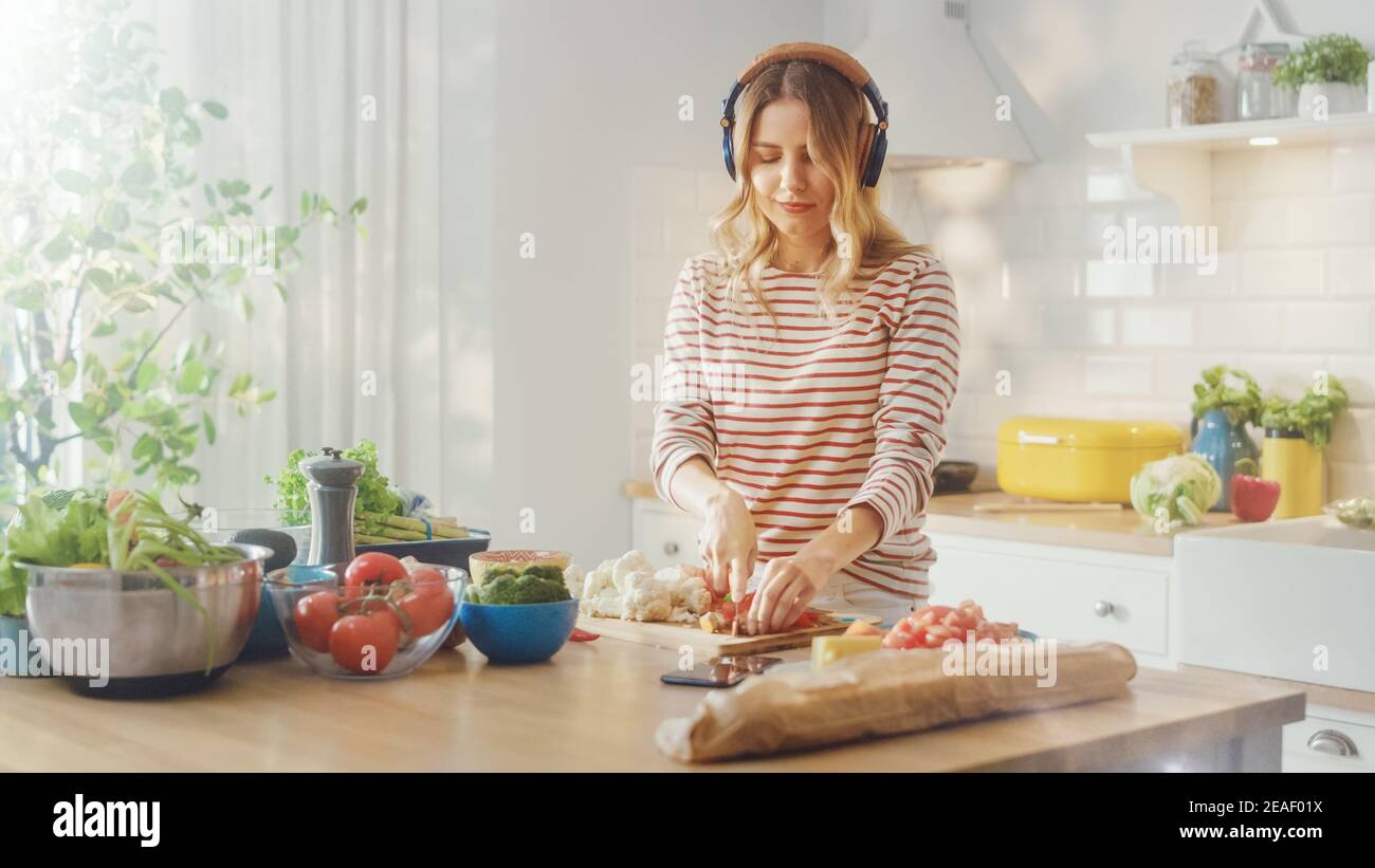 Beautiful Young Female Preparing a Healthy Organic Salad Meal in a Modern Sunny Kitchen. She Wears Headphones and Dances to the Music from a Mobile Stock Photo
