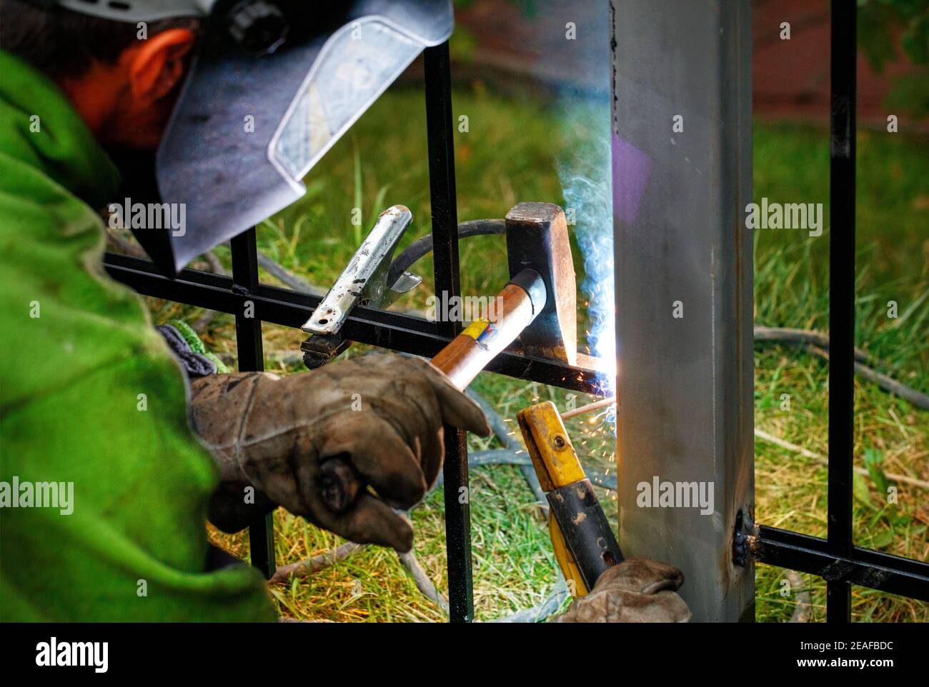 A welder wearing a safety helmet and gloves is welding a metal fence, bright sparks fly, blue smoke, selective focus, copy space. Stock Photo