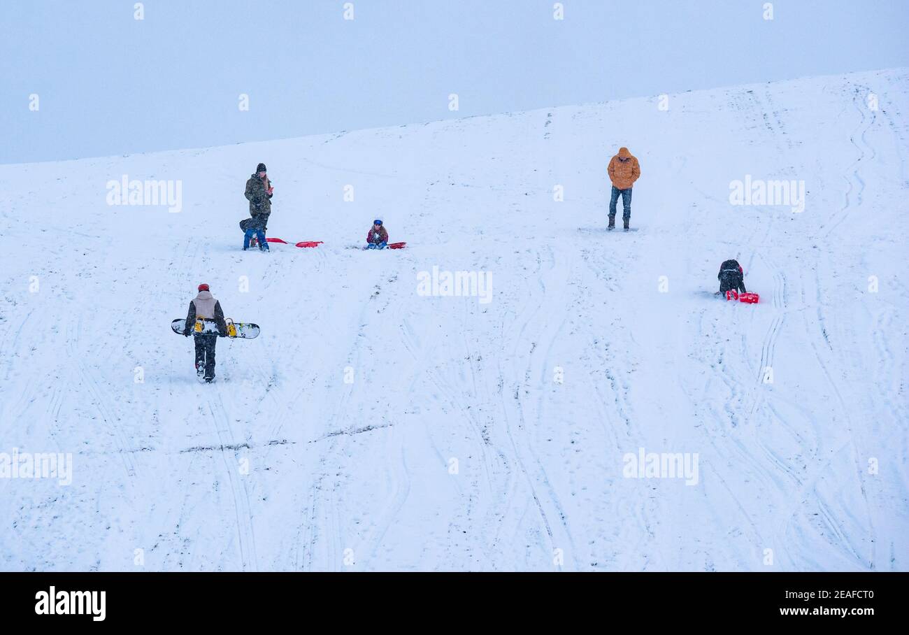 East Lothian, Scotland, United Kingdom, 9th February 2021. UK Weather: sledging on Skid Hill. People enjoy the Winter snow on the appropriately named Skid Hill. A family carry sledges up the hill as a man walks up with a snowboard Stock Photo
