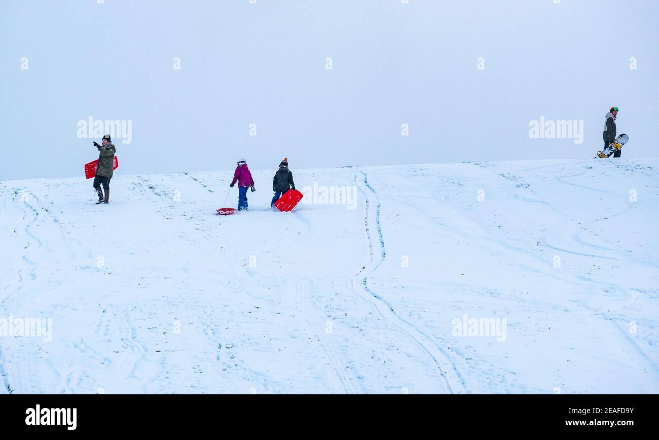 East Lothian, Scotland, United Kingdom, 9th February 2021. UK Weather: sledging on Skid Hill. People enjoy the Winter snow on the appropriately named Skid Hill. A family pull their sledges to the top of the slope as a man carries a snowboard Stock Photo