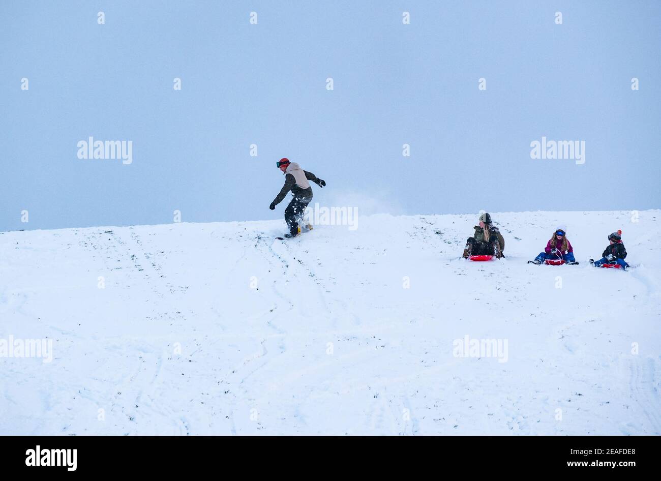 East Lothian, Scotland, United Kingdom, 9th February 2021. UK Weather: sledging on Skid Hill. People enjoy the Winter snow on the appropriately named Skid Hill. A family on the slope on sledges with a man snowboarding Stock Photo