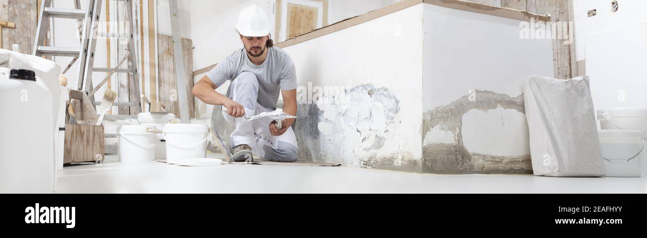 plasterer man at work, take the mortar from the bucket with trowel to plastering the wall of interior construction house site and wear helmet, panoram Stock Photo