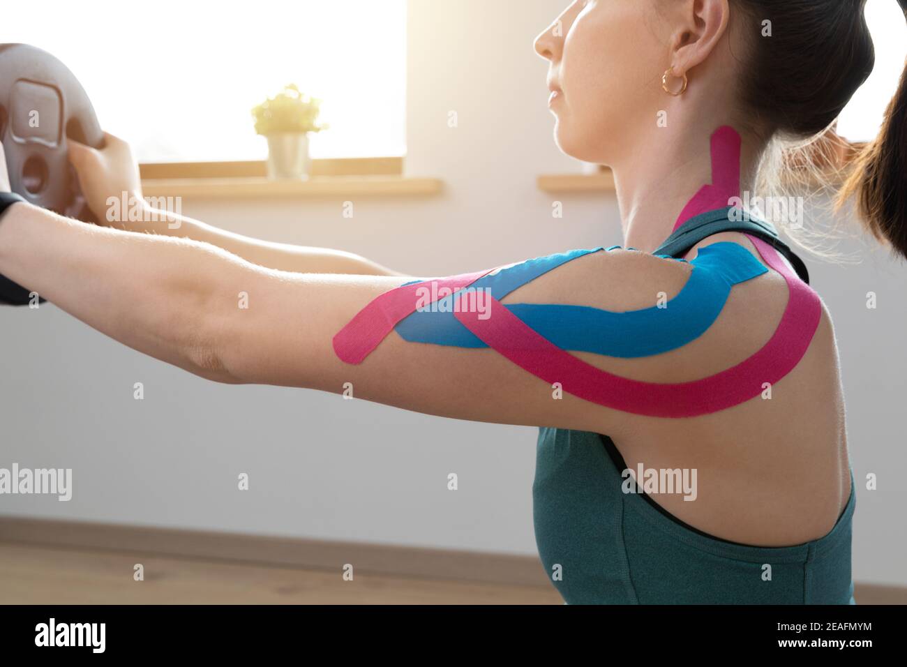 Young fit women performing excersise with elastic therapeutic kinetic tape on her shoulder on a sunny day at home. Kinesiology physical therapy. Stock Photo