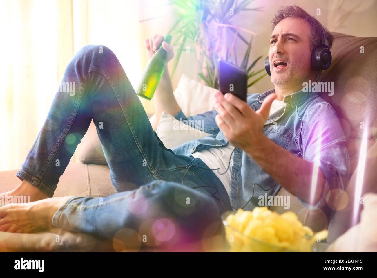 Happy man singing on the sofa at home taking a snack on a sofa while listening to music with headphones from a mobile Stock Photo