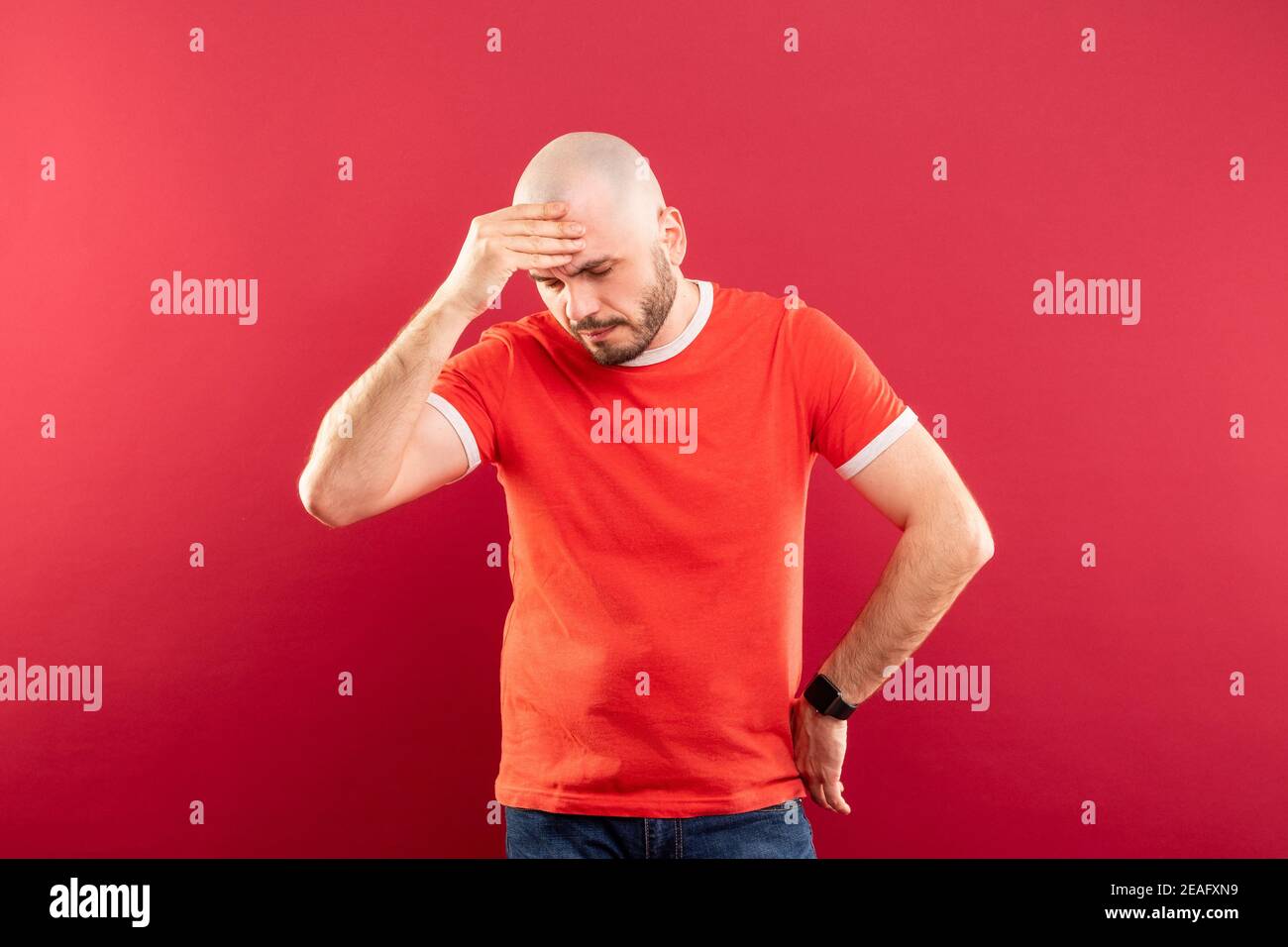 A bald middle-aged man with a beard and in a red T-shirt on a red background. Holds his head with his hands. He has a headache. Stock Photo