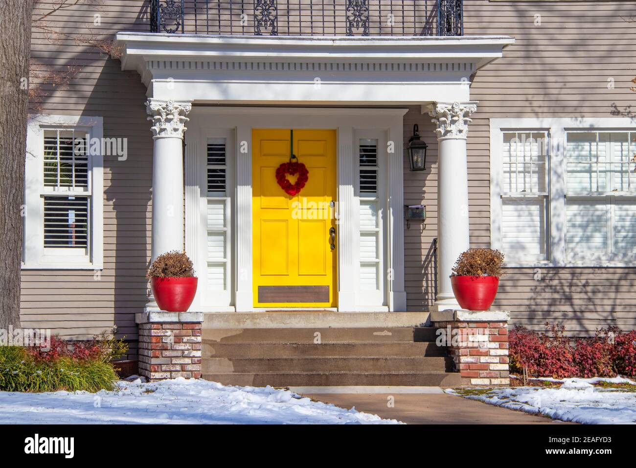 Entrance of pretty vintage house with ornate columns on porch and red valentine wreath on bright yellow door in snow Stock Photo
