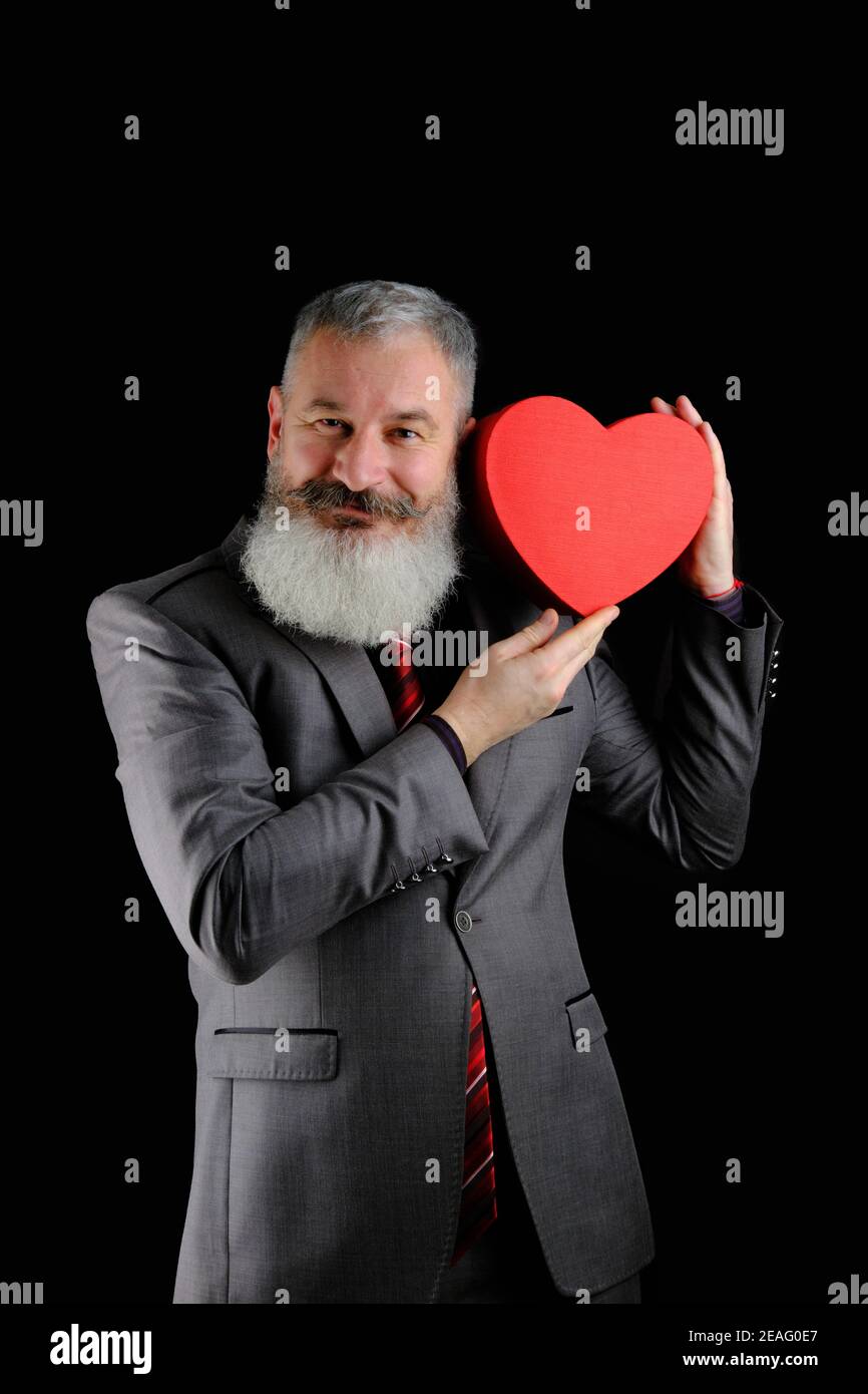 Mature bearded man wear gray suit holds red heart shaped gift box, isolated black background Stock Photo