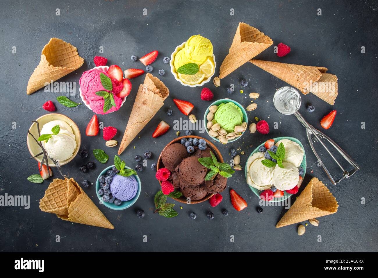Various colorful ice cream balls in different bowls, with ice cream waffles cones and flavor ingredients - pistachio nuts, berries, lemon, chocolates, Stock Photo