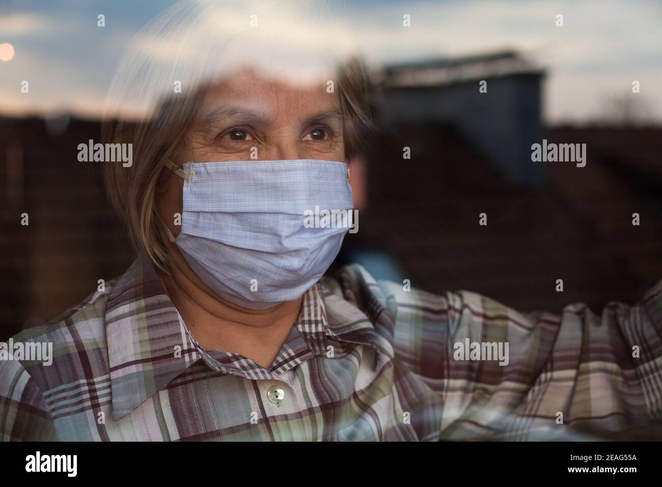 Elderly caucasian woman wearing handmade protective face mask,nursing care home,looking outside the window with sadness in her eyes,self isolation Stock Photo