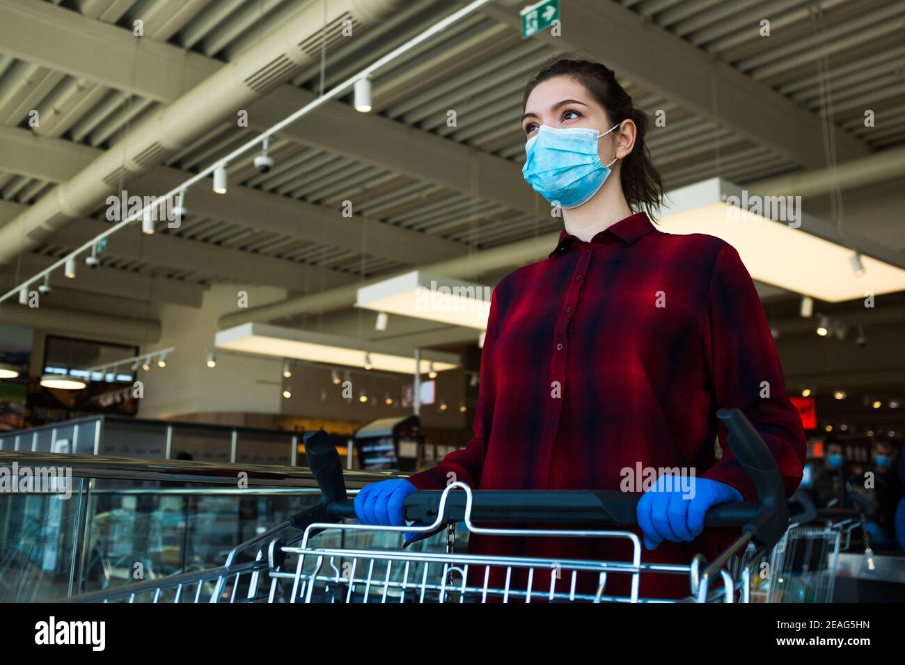 Young caucasian woman pushing shopping cart in supermarket, wearing protective blue surgical face mask and latex gloves, health and safety measurement Stock Photo