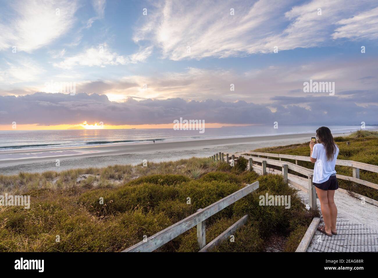 Young women taking pictures with a mobile phone of sunrise at Ruakaka Beach, New Zealand Stock Photo