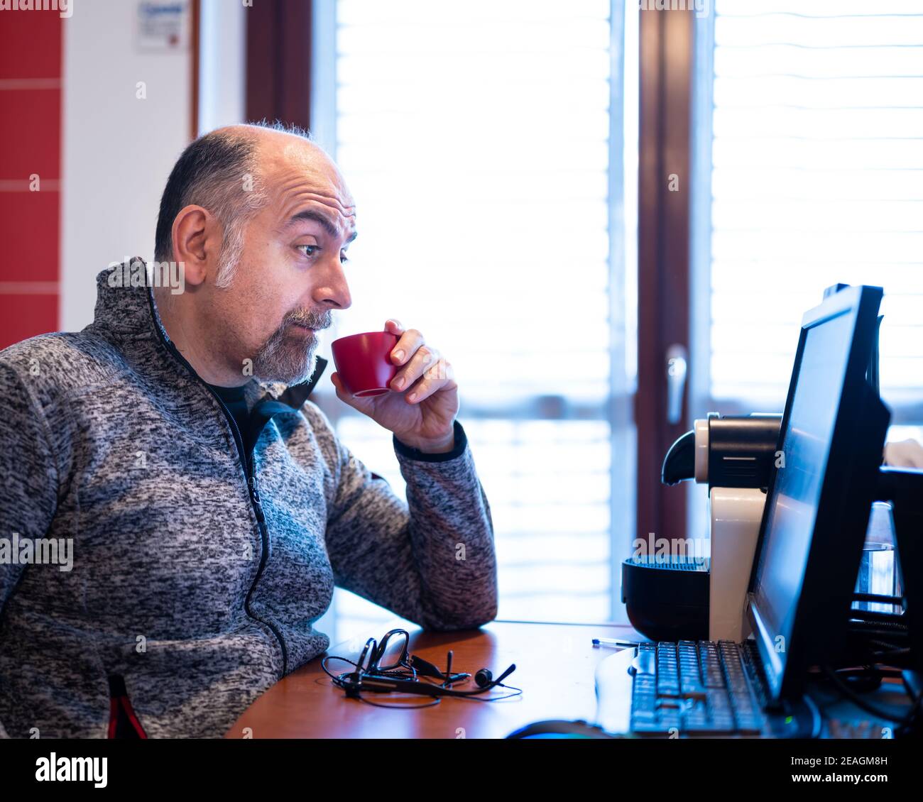 A middle aged caucasian man working from home, while drinking a coffee, is pleasantly surprised by a notification that reads on his computer monitor. Stock Photo