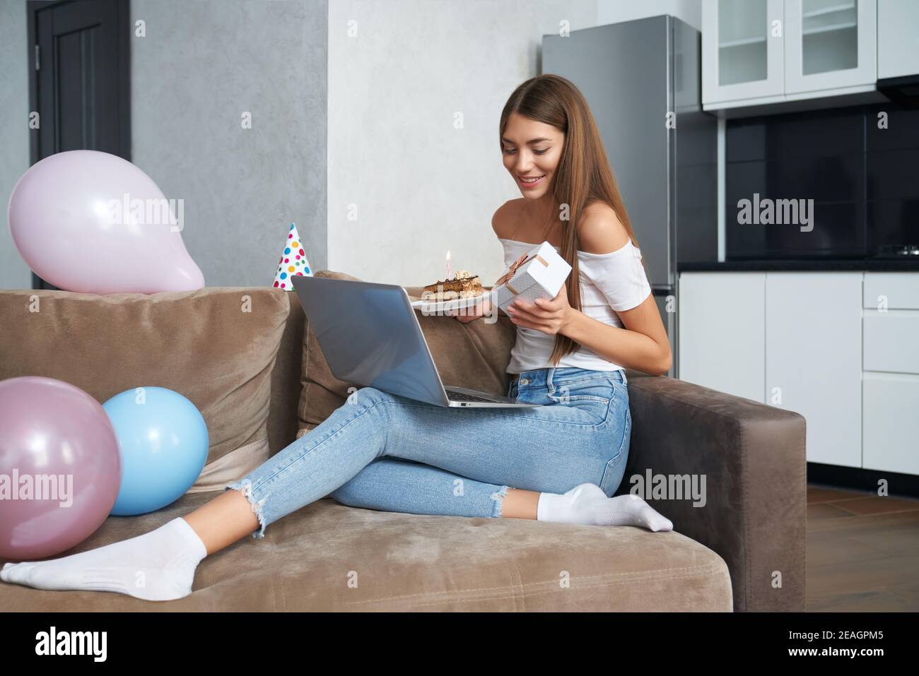 Beautiful young lady sitting on couch with laptop on knees and holding cake with candle and gift box. Woman having online celebration of her birthday Stock Photo