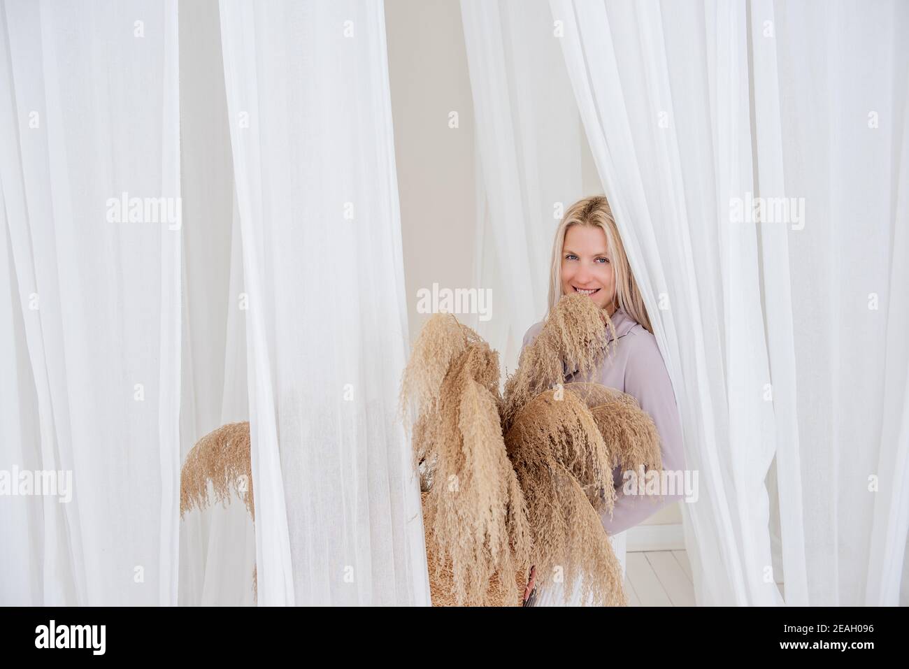 Young blonde woman in a lilac shirt, white dress, holds in her hands straw basket with pampas grass among the airy, developing tulle fabrics. Minimali Stock Photo