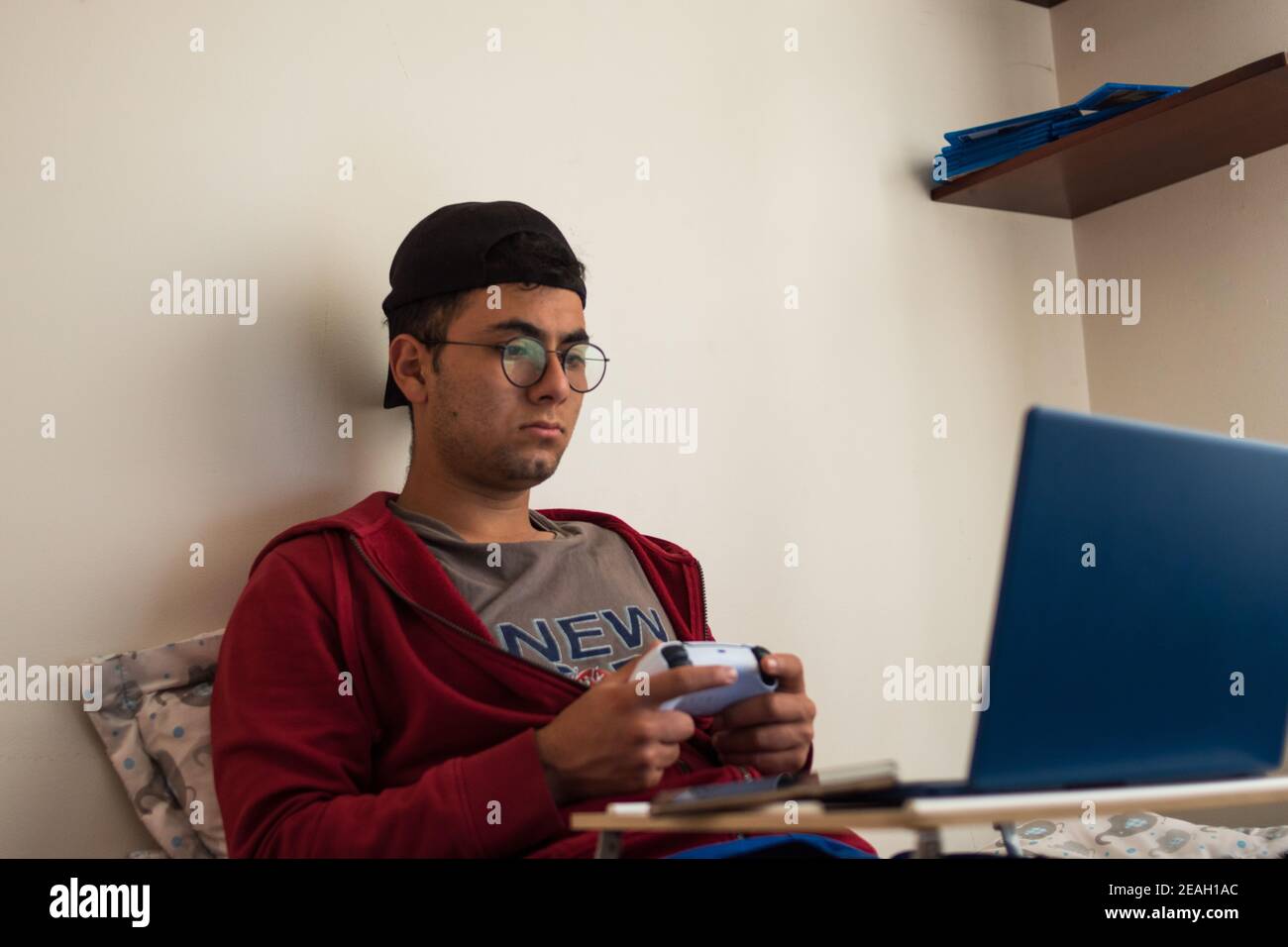 Young man wearing a red jacket playing videogames with a white controller on his blue laptop at home Stock Photo