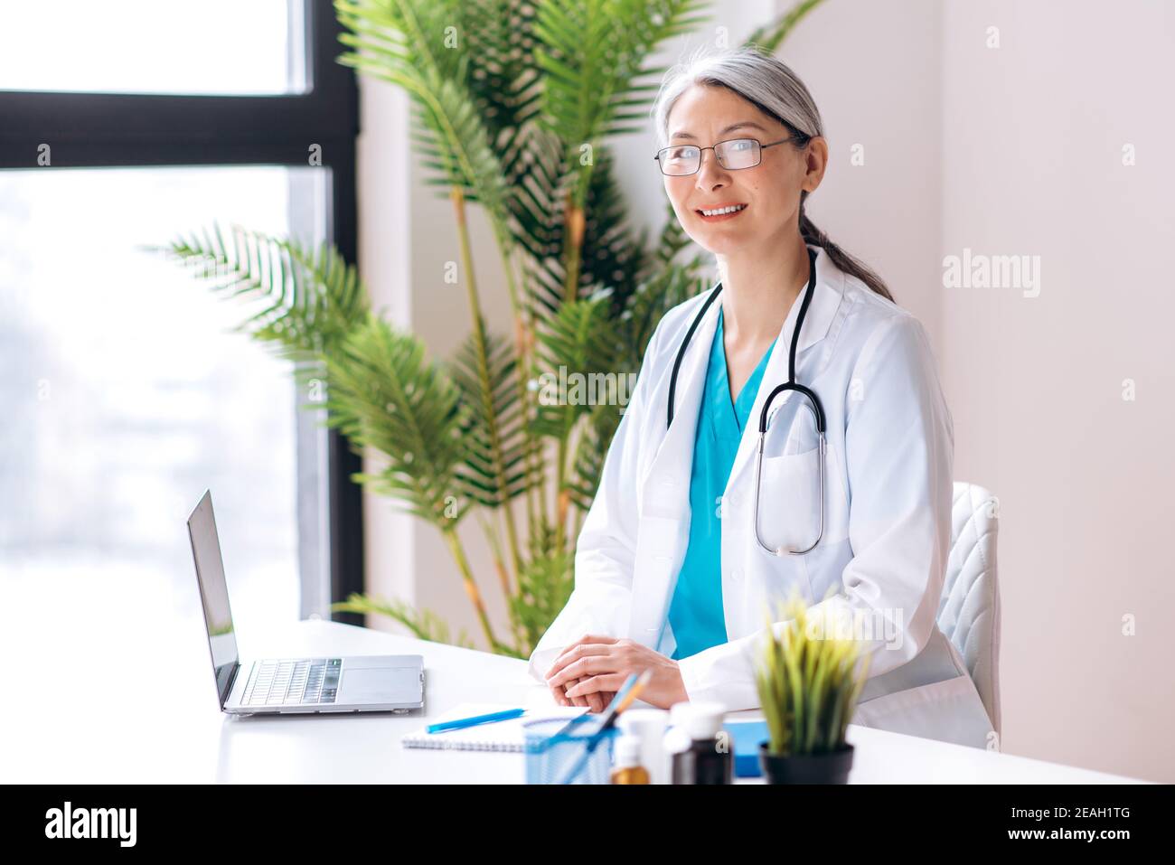 Gray-haired female middle aged general doctor dressed in medical uniform and eyeglasses looks directly at the camera at workplace at the clinic. Medical help and consultation, health concept Stock Photo