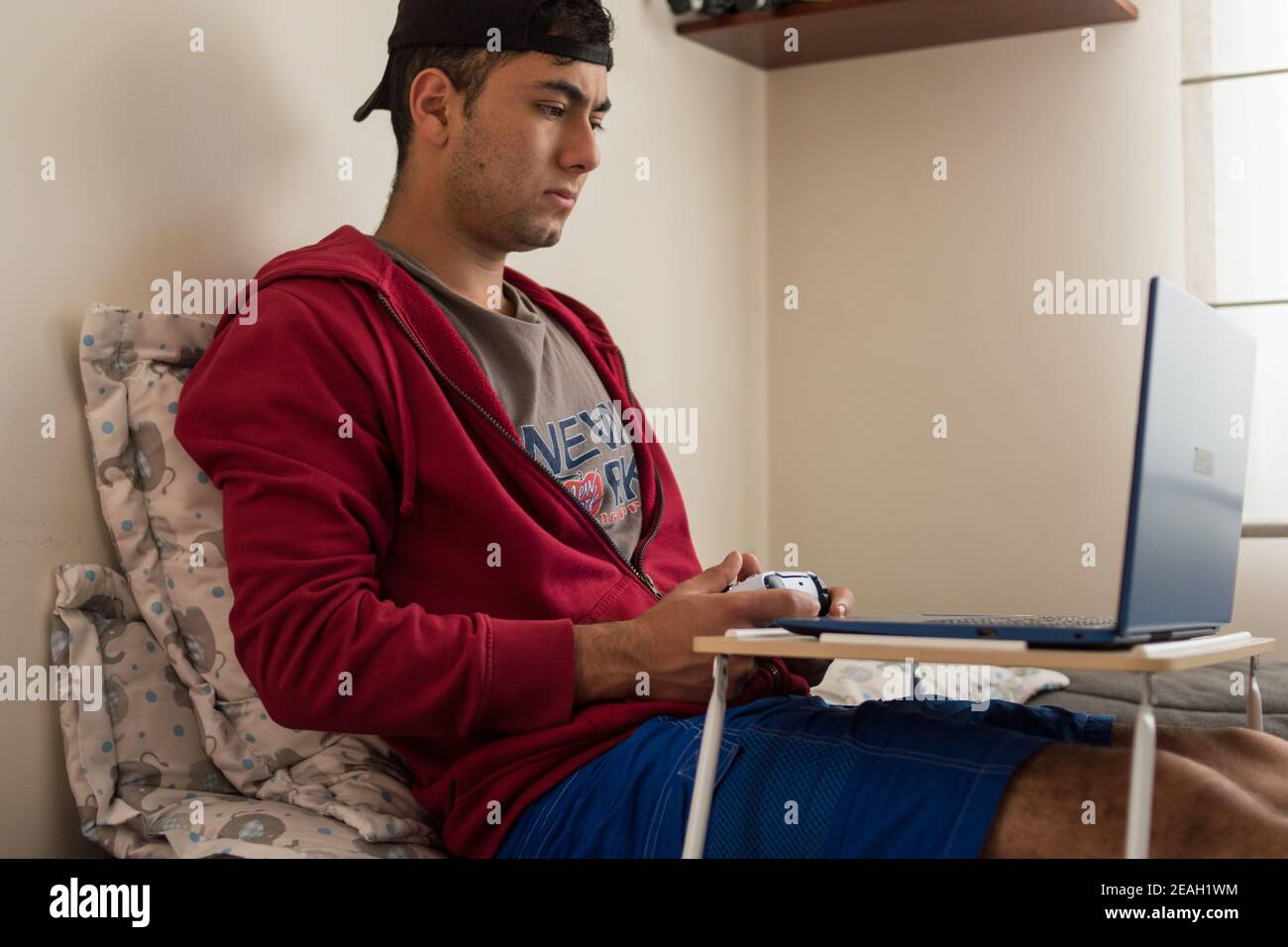 Young man wearing a red jacket playing videogames with a white controller on his blue laptop at home Stock Photo
