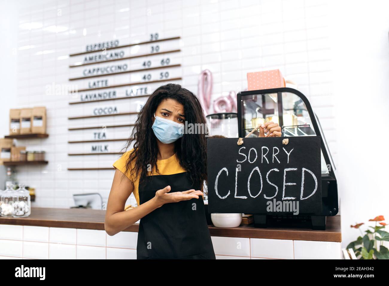 Sad African American waitress, barista or small business owner. Unhappy young woman in a black apron and medical mask, stands inside coffee shop, restaurant or bar and holds a CLOSED sign board Stock Photo