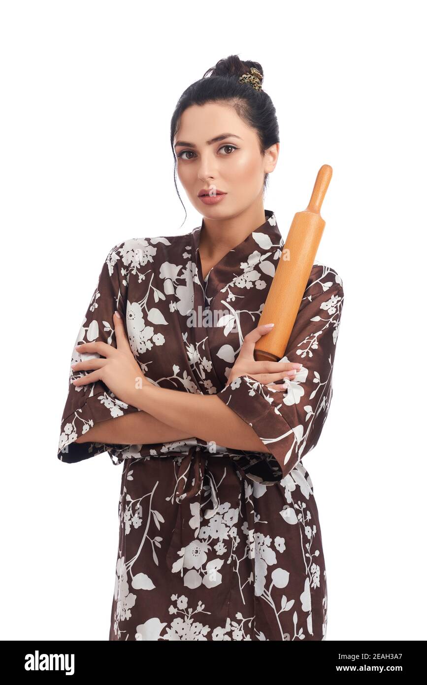 Front view of beautiful young woman wearing domestic robe, holding rolling pin and looking at camera. isolated over white studio background. Concept o Stock Photo