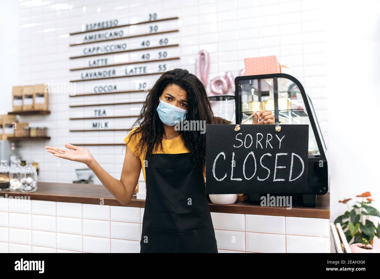 Unhappy sad African American waitress, barista or small business owner in a black apron, stands inside coffee shop, restaurant or bar holds a signboard CLOSED Stock Photo