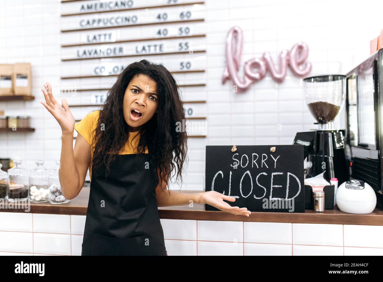 Frustrated emotional African American waitress, barista or small business owner in a black apron, stands inside coffee shop, restaurant or bar near a signboard CLOSED Stock Photo