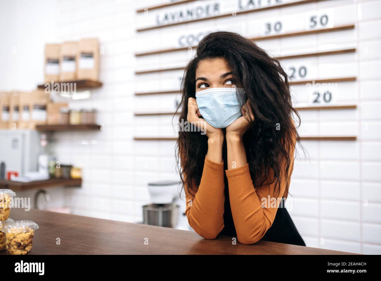 Sad young African American female barista wearing uniform and protective medical mask stands behind the bar counter of coffee shop and waiting for clients. Coffee bar concept Stock Photo