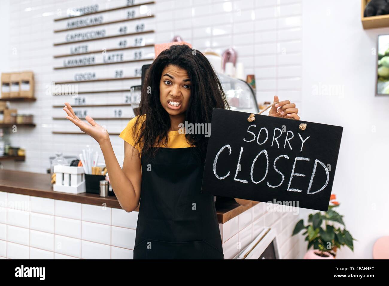 Unhappy emotional black waitress, barista or small business owner in a black apron, stands inside coffee shop, restaurant or bar near a signboard CLOSED Stock Photo