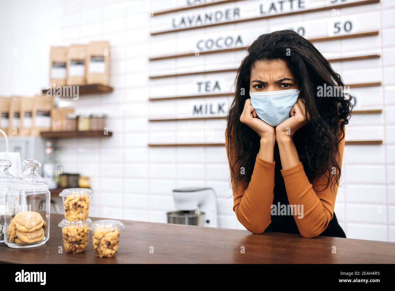 Frustrated waitress wearing uniform and protective medical mask. Young African American female barista is sad because of quarantine, stands behind the bar counter of coffee shop. Small business Stock Photo