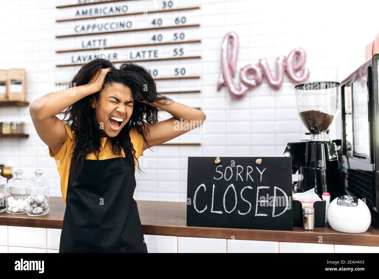 Emotional frustrated African American waitress, barista or small business owner in a black apron, stands inside cafe, restaurant or bar near a signboard CLOSED, screams and holds his head with hands Stock Photo