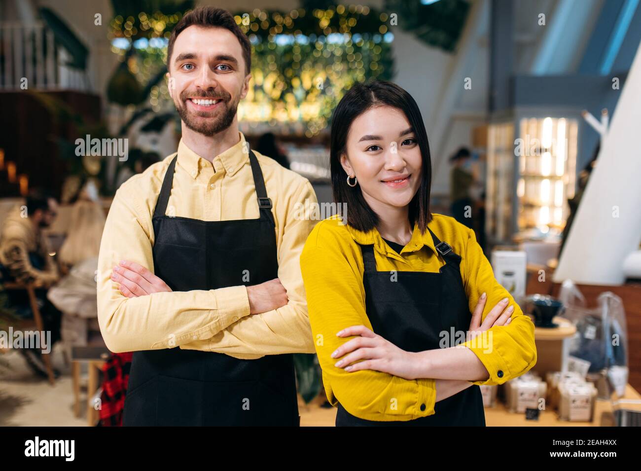 Portrait of two friendly waiters. Attractive caucasian guy and beautiful asian girl in black aprons stand with arms crossed inside a restaurant, cafe or bar and smiling. Teamwork concept Stock Photo