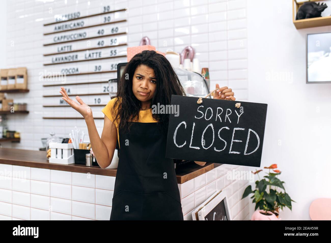 Frustrated sad multiracial waitress, barista or small business owner in a black apron, stands inside coffee shop, restaurant or bar, holds a signboard CLOSED with confused face expression Stock Photo