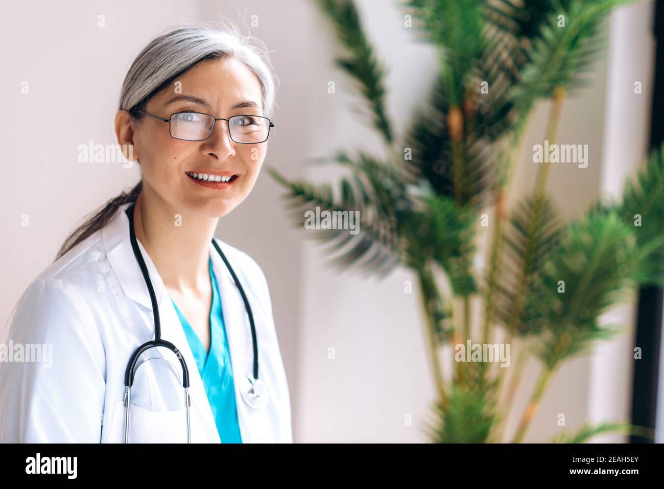 Portrait of gray-haired female middle aged doctor dressed in medical uniform and eyeglasses looks directly at the camera at workplace at the clinic and smile. Medical help and consultation, health Stock Photo